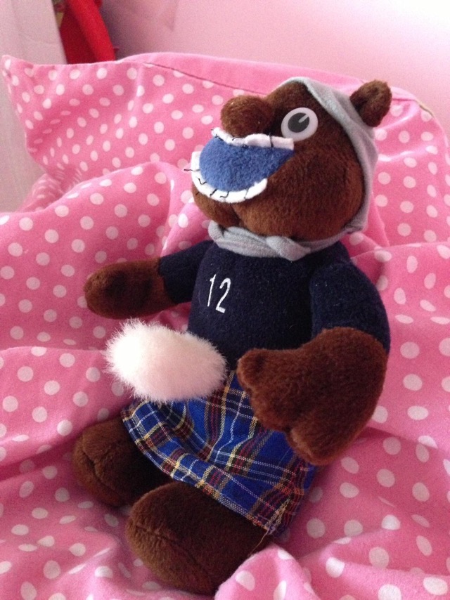 Shuggie Bear, one-time mascot of the Scottish Claymores American Football team (NFL Europe). He's wearing a badly-fitting grey cloth helmet, a blue jersey with white nunber 12, a white sporran extending out from his midriff in a slightly disturbing manner, and a predominantly blue tartan kilt. He's photographed on a pink pillow with pale pink spots.