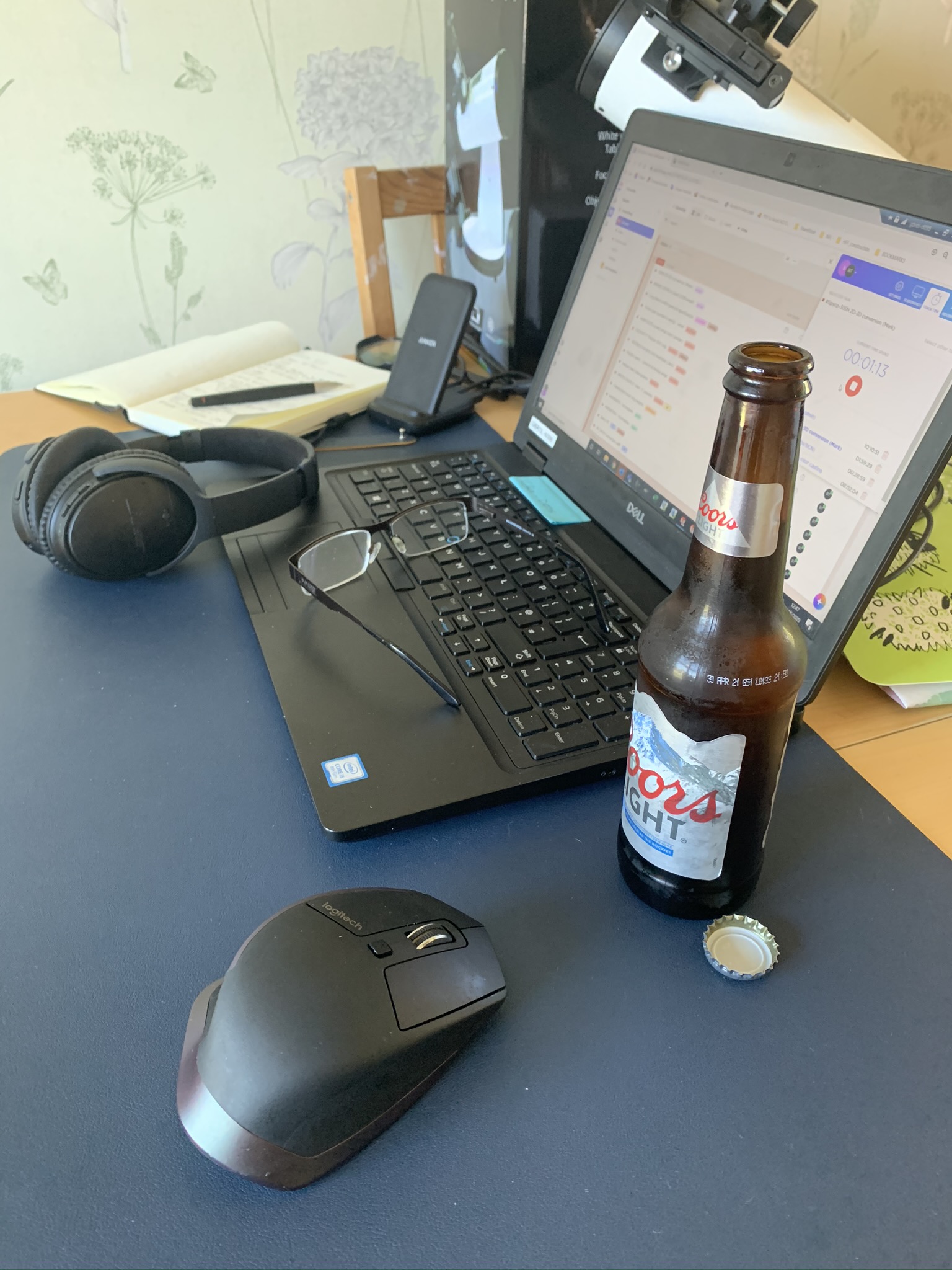 A dining room table repurposed into a working desk. Laptop, mouse, headphones, reading glasses, phone stand, notebook, 4-colour pen, opened beer, telescope…