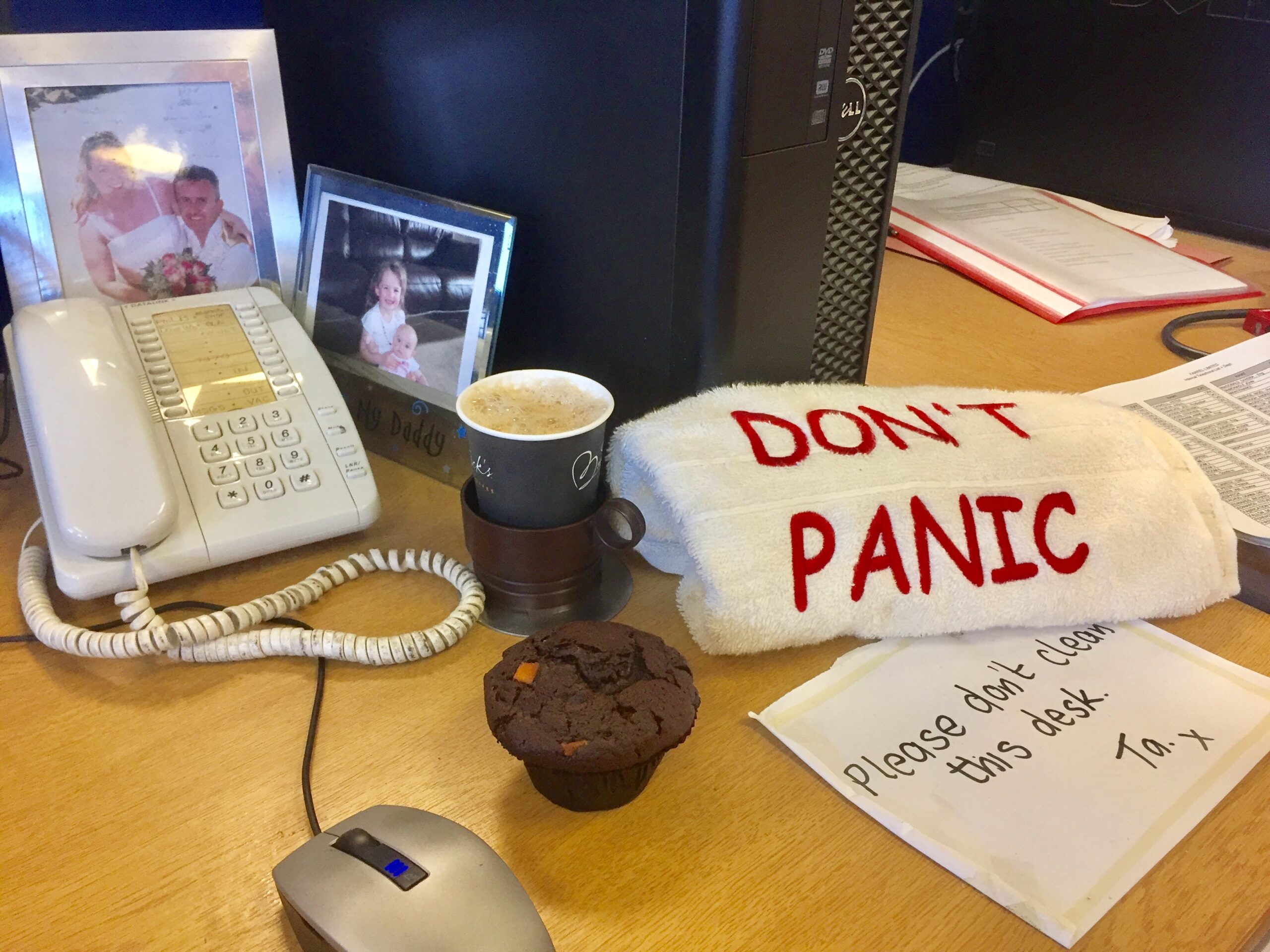 A 2017 photo of a work desk with old-fashioned phone, simple mouse, chocolate muffin, plastic cup, family photos, computer system unit, pieces of paper, and a folded towel embroidered with the words "DON'T PANIC".