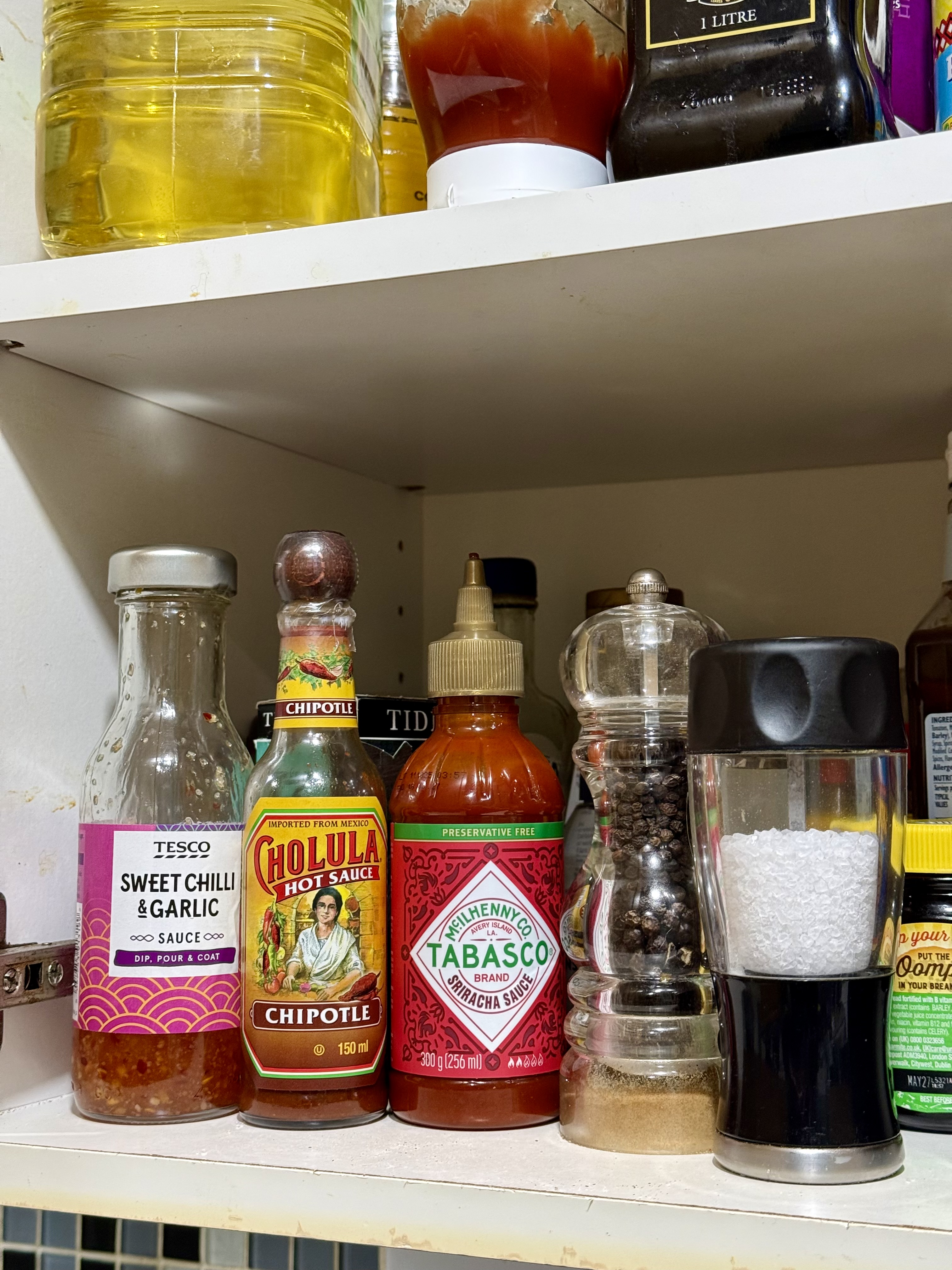 A cupboard full of sauces, condiments, cans and stuff to be cooked. Of particular interest are the sweet chilli and garlic sauce, chipotle sauce and Tabasco & sriracha sauce bottles.