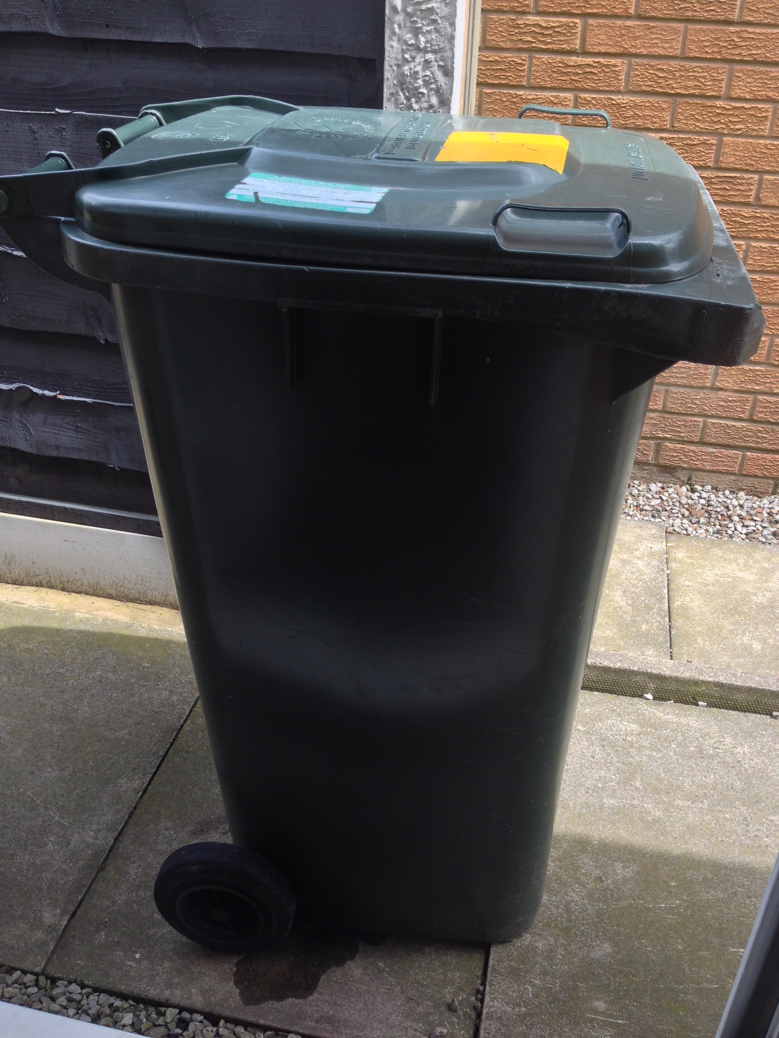 A dark green UK wheelie bin on paving slabs.