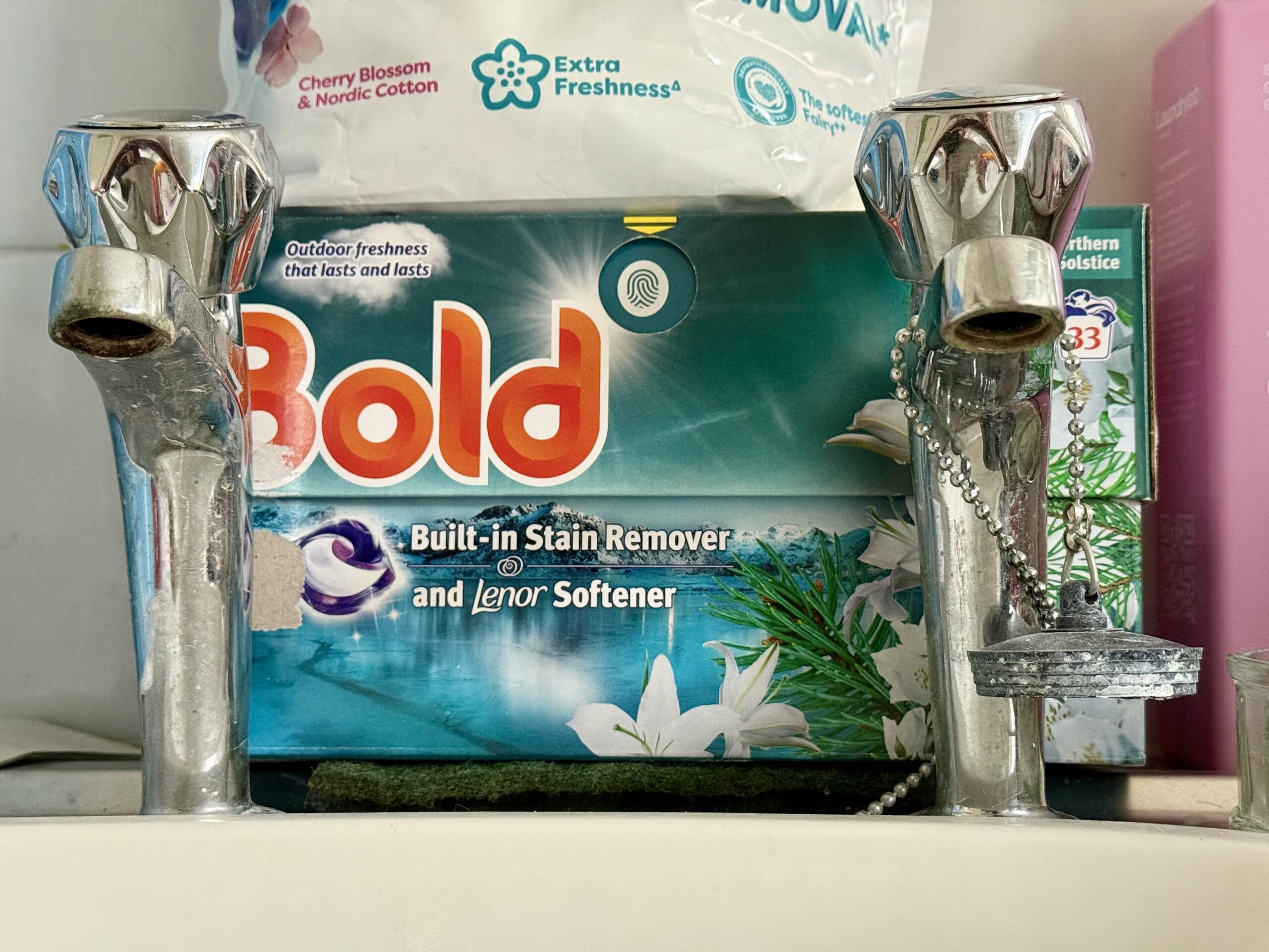 A pair of taps, left-cold, right-hot, with a box of Bold washing machine capsules behind and a pack of Fairy Big One capsules on top of that. Chances are these taps have been in the house since it was built in 1988. We have no sentimental attachment to them.