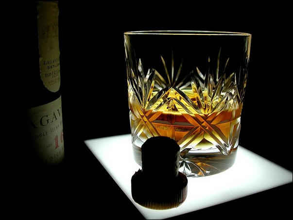 A whisky glass sat on a small illuminated light table. There's a silhouetted cork in the foreground and a dimly-lit bottle on the left background. The bottle contains a Lagavulin 16 year old whisky, as does the glass. The photo is high contrast.